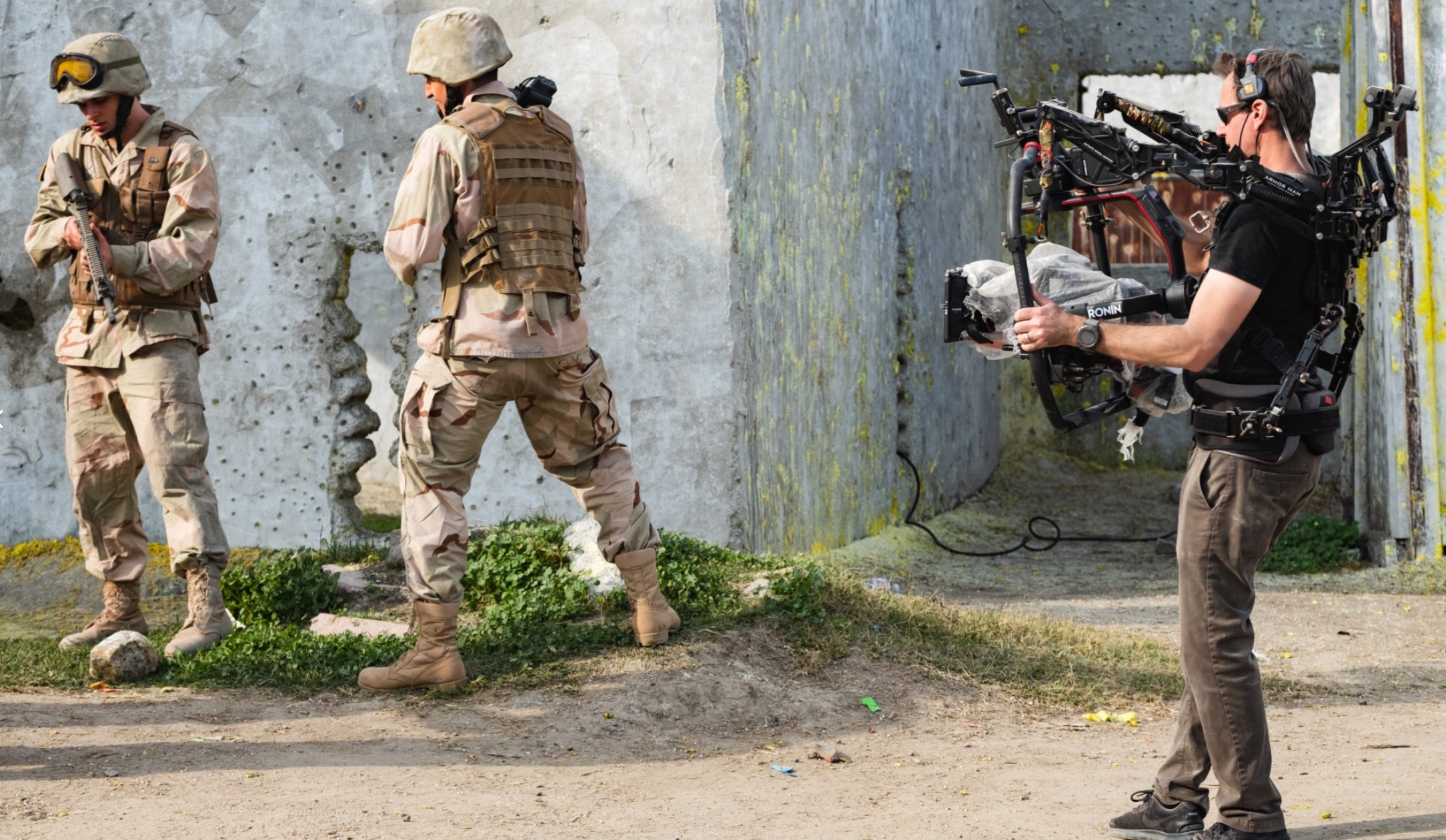 屋外の撮影現場。右側には、黒い外骨格のようなサポートシステム「Tilta Armor Man 3.0」を身体に装着したカメラマンが、大きなジンバルに載ったカメラを構えて歩いている。左側の奥には、砂漠迷彩の軍服とヘルメットを着用し、銃を構えた2人の兵士役のキャストが、コンクリートの壁のそばで演技をしている。