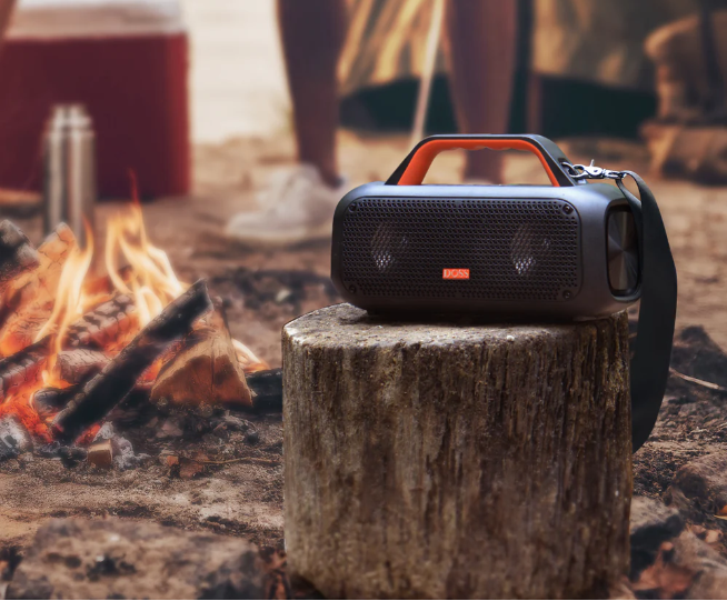 焚き火のそばの切り株に置かれたスピーカーと、背後にキャンプ道具や人物の足元が見える屋外使用のシーン