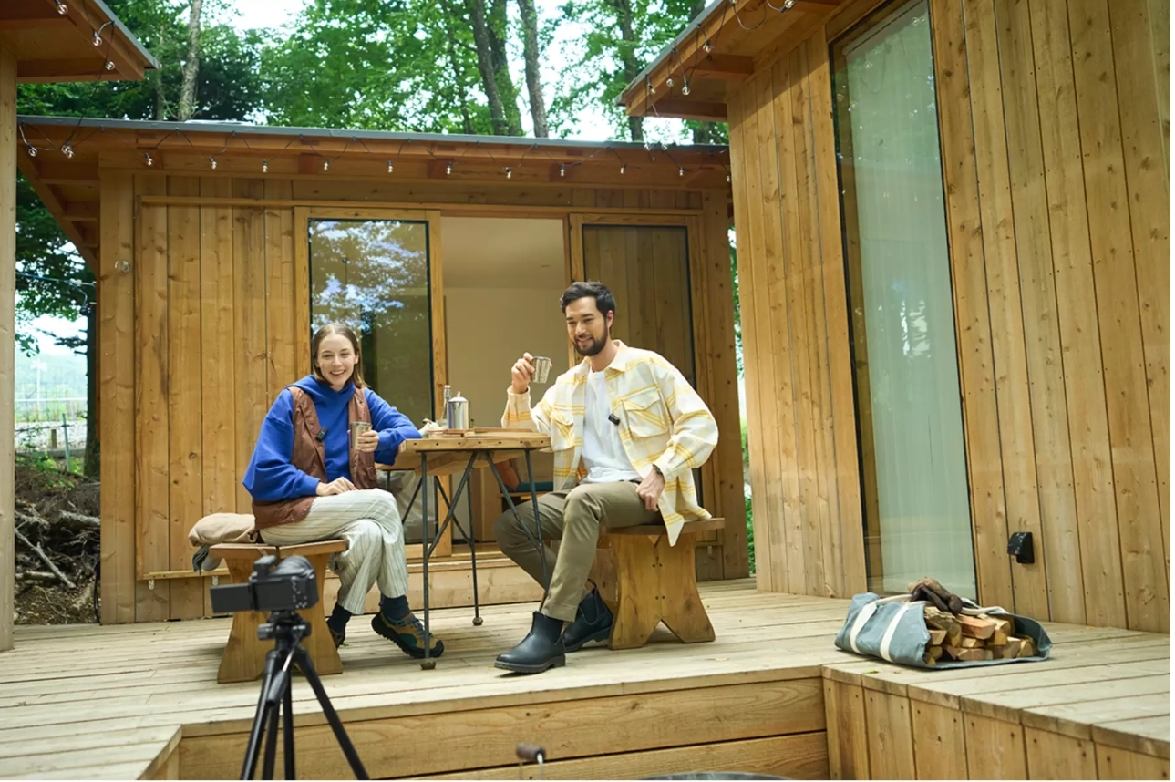 木造の小屋を背に、三脚に据えられたカメラの前で、テーブルを囲み飲み物を手に談笑する男女のVlog撮影風景。